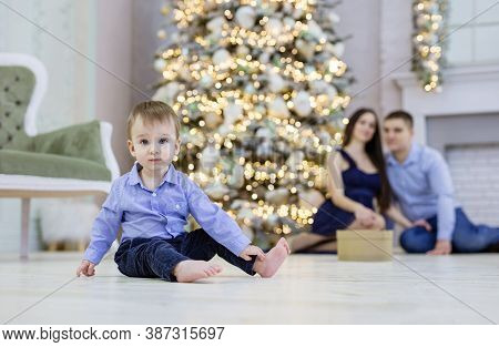 Cute Baby Sitting On Floor And Looking At Carema. His Parents Watching Him At Christmas Tree. Shallo