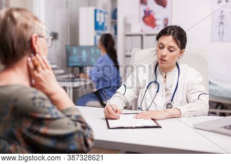 Doctor In Hospital Office Writing Treatment For Senior Woman During Examination. Nurse Wearing Blue 