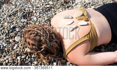 A Young Woman Is Lying On A Pebble Beach. Concept. Man Hand Putting Hot Stones On The Back Of A Youn