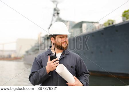 Maritime Assistant Engineer Talking By Walkie Talkie, Holding Blueprints And Standing Near Vessel.