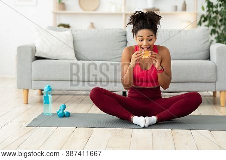 Hungry African American Lady Eating Burger Having Cheat Meal After Fitness Workout Sitting On Mat At