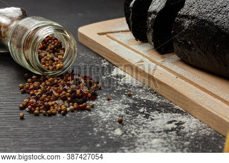 fresh black rye bread on a cutting board with seasonings sprinkled with flour on a black wooden tabl