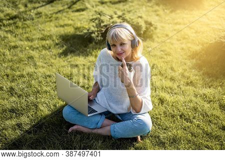 Adult Woman Texting Laptop, Sitting On The Grass In Headphones Outside In Park. Working And Drinking