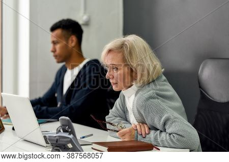 Aged Woman, Senior Intern Looking Focused At The Screen While Using Laptop, Sitting At Desk, Working