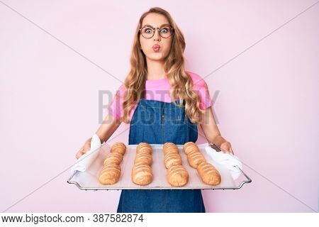 Young caucasian woman with blond hair wearing baker uniform holding homemade bread puffing cheeks with funny face. mouth inflated with air, catching air. 