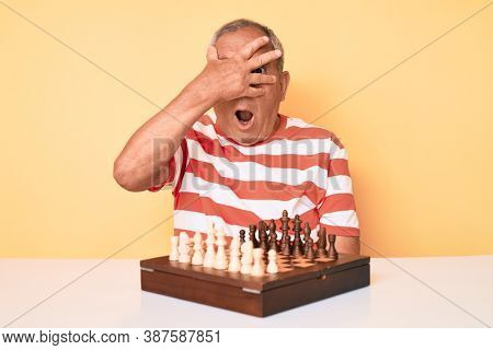 Senior handsome man with gray hair playing chess sitting on the table peeking in shock covering face and eyes with hand, looking through fingers afraid 
