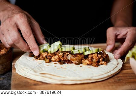 closeup of a young caucasian man preparing a durum or a burrito, with chicken meat cooked with different vegetables such as onion or red and green pepper, and fresh lettuce and raw cucumber