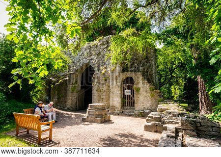Visby, Sweden - July 27, 2020: A Senior Couple Sitting On A Bench Next To The Entrance Of An Ancient