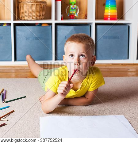A Cute Blond Boy In A Yellow T Shirt Is Lying On The Floor With A Sheet Of Paper And Pencils And Loo