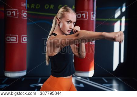 Serious Blond Female Boxer Punching An Air In Empty Modern Gym With Two Red Punching Bags In Backgro