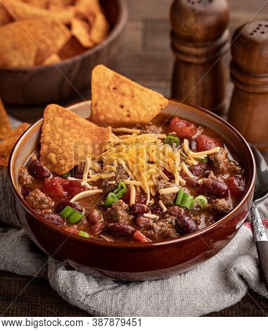 Bowl Of Chili Con Carne With Shredded Ceddar Cheese And Nacho Chips On A Rustic Wooden Table.