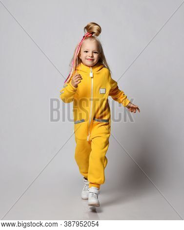 Full-growth Portrait Of Joyful Smiling Blonde Kid Girl With Colorful Braids Wearing Modern Yellow Ju