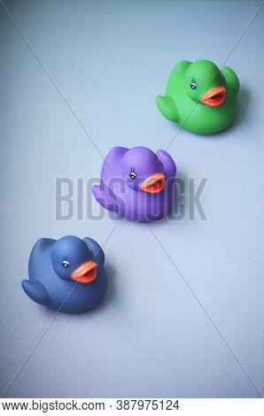 Three Isolated Toy Rubber Ducks In Dark Blue, Green And Violet Color On A Grey Background