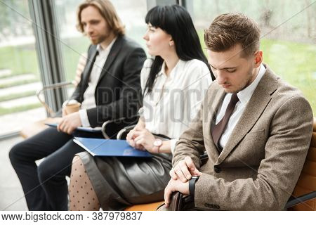 Three Office Workers Are Waitig For An Appointment. The Man In The Right Is Looking At His Watch