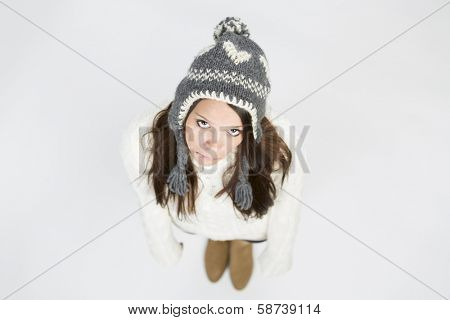 Upset young woman in warm winter clothing and grey cap, standing with arms hanging down and looking up with sadness, isolated on grey background.