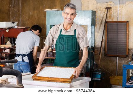 Portrait of smiling male worker dipping mold in pulpy water while coworker working at paper factory