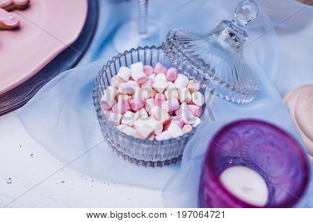 Colorful mini marshmallows in Glassware on a white table with blue lace. Different mini white and pink puffy marshmallows. Marshmallow concept. Selective focus