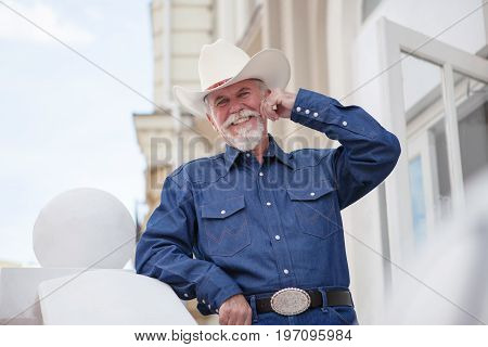 A mature cowboy in a hat, jeans and a denim shirt looks at the camera. On open air