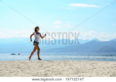 Active sporty woman run along ocean surf by water pool. Sunset sand beach background with blue sky and sun. Woman fitness, jogging workout and sport activity on summer holiday.