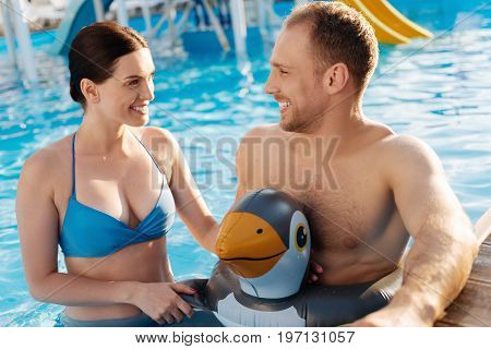 Quality time. Pleasant young man in a bird-shaped swim ring and his charming wife standing waist-deep in the swimming pool and talking to each other merrily