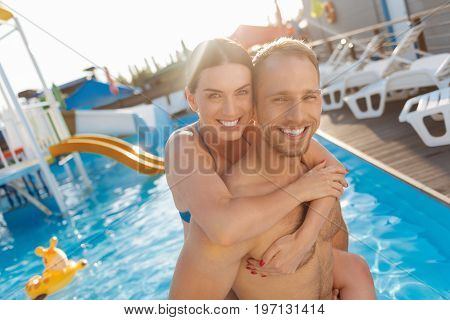 Sweet hug. Adorable young woman hugging her husband from behind while both of them standing in the swimming pool and happily smiling at the camera