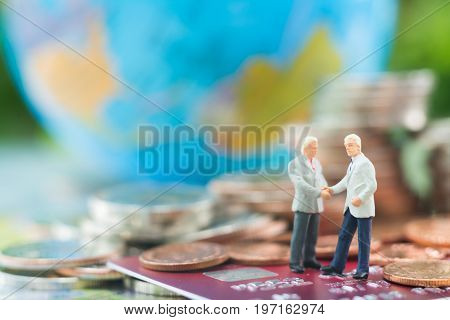 Miniature businessman handshaking and standing on the credit card or debit card with blurred globe and stacks of coins background using as commitment agreement investment partnership E-commerce and online business concept.