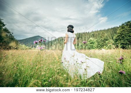Beautiful young woman wearing elegant white dress standing on a feild and looking to the mountains