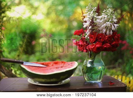 water melon cut with knife and roses flowers in vase on green summer garden background
