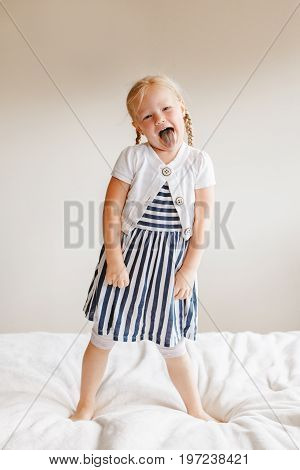 Portrait of cute finny white Caucasian blonde girl jumping on bed at home and showing tongue. Hilarious active kid having fun indoors. Authentic lifestyle childhood concept.