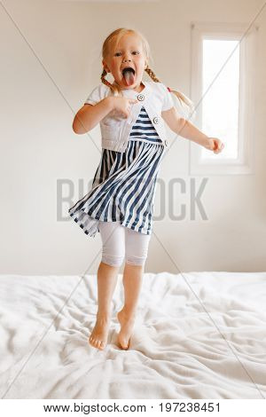 Portrait of cute finny white Caucasian blonde girl jumping on bed at home and showing tongue. Hilarious active kid having fun indoors. Authentic lifestyle childhood concept.