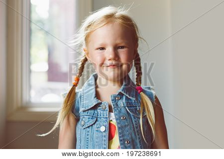 Closeup portrait of cute adorable white blonde Caucasian smiling girl looking in camera. Child girl with light fair hair with plaits wearing country village outfit jeans vest. Toned with filters.