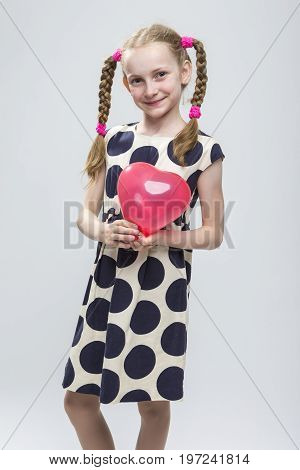 Portrait of Funny Caucasian Blond Girl With Pigtails Posing in Polka Dot Dress Against White. Holding Red Air Balloon. Vertical Shot
