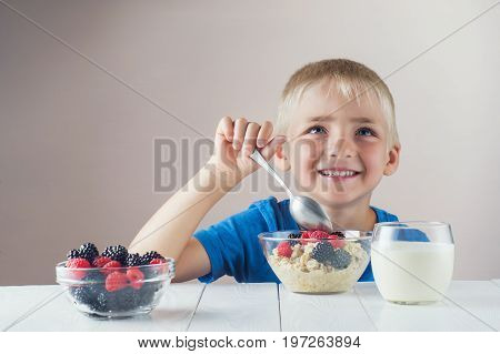 Happy smiling child eating oatmeal with berries. The concept of a healthy breakfast for children porridge with raspberries and blackberries a glass of yogurt on white wooden table.