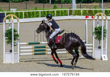 Young girl jumping with black horse. Blond pretty little girl going to jump a hurdle in a competition. Girl with horse during equestrian show jumping. Bright sunny day.