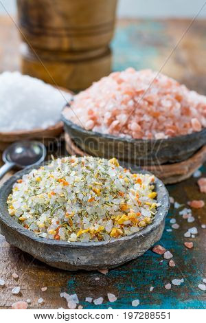 Different types of natural salt in stone bowls on wooden surface. White sea salt pink Himalayan salt Spiced salt with rosemary