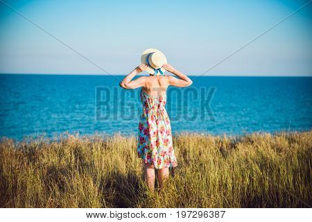 Summer Portrait Of Young Pretty Woman In A Straw Hat And Dress Standing Backwards In The Grass And L