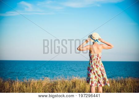 Summer Portrait Of Young Pretty Woman In A Straw Hat And Dress Standing Backwards In The Grass And L