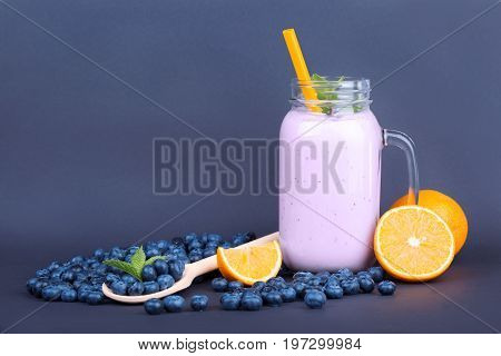 Close-up of a glass jar with bilberry smoothie on a dark blue background. A bunch of bilberries and cut oranges are near a full jar of bilberry drink with orange straw and decorative mint.
