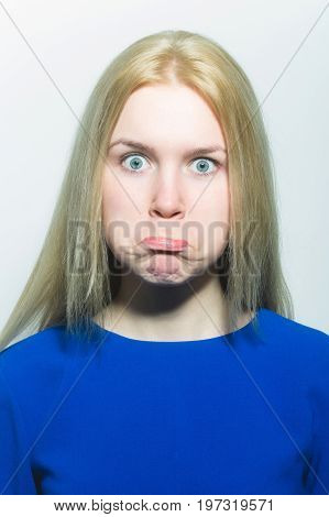 Girl puffing cheeks on funny face. Young woman with crossed grey eyes healthy skin and long blond hair in blue dress isolated on white. Grimace and facial expression. Strabismus