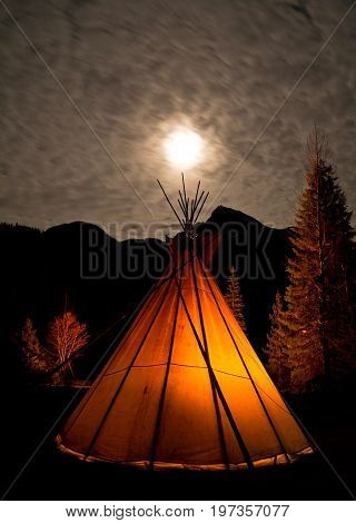 A teepee illuminated by a campfire from within under the full moon.