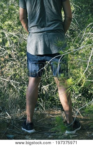 a young caucasian man wearing shorts and T-shirt seen from behind peeing in the woods