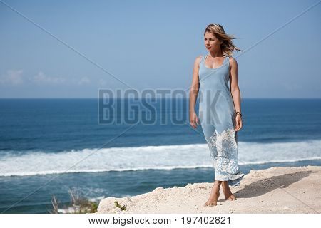 Beautiful girl with hair floating in the air and light-blue dress is standing on the rock ledge in front of the ocean and looking away