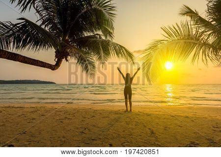 Silhouette of young beautiful girl raised hands on tropical beach at sunset