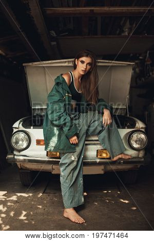 Girl worker in dirty green overalls stands in workshop near opened capote of retro car.