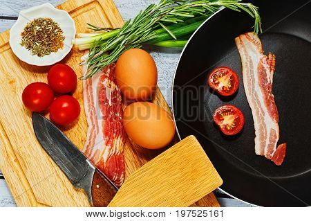 Rural style concept pf omelet or fried eggs preparation with pan different ingredients including pork bacon cherry tomatoes and herbs and spices mix