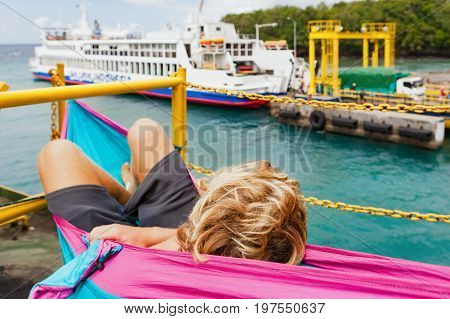 Backpacker traveling lifestyle. Passenger relaxing in hammock on ship deсk looking at public ferry. Cars landing on board at cargo pier in Bali island harbour Padang bay to cross Lombok straits.