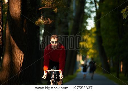 Candid shot of young hipster with bristle cycling along alley in public park. Trendy looking student enjoying ride on his fixed gear bike commuting home from college. Hobby and active lifestyle