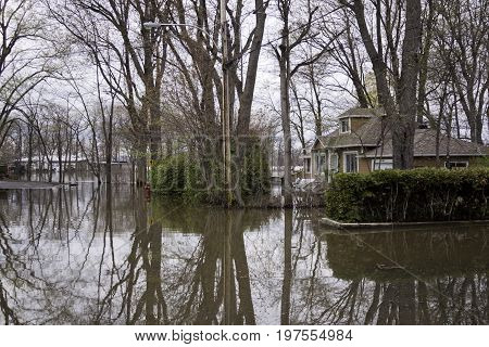 Close up landscape view of flood waters rising over a paved road in front of properties with trees foliage and the houses partly submerged in the distance in the West Island of Montreal, Quebec on a bright overcast day in May.