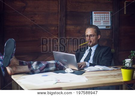 Freelance wearing business jacket, shirt and pajamas trousers. Man at home office holding legs on wooden desk while working with laptop. Toned image.