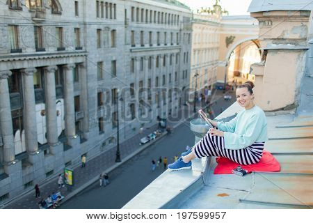 Attractive And Stylish Girl Sits On The Roof Of The House In The Old Town And Reads The E-book